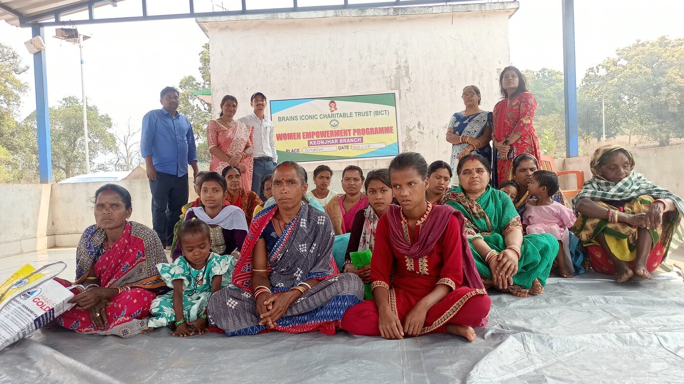 Women and children from a tribal community in Keonjhar district gather beneath a Brains Iconic Charitable Trust Women Empowerment Programme banner in Dumuridihi.