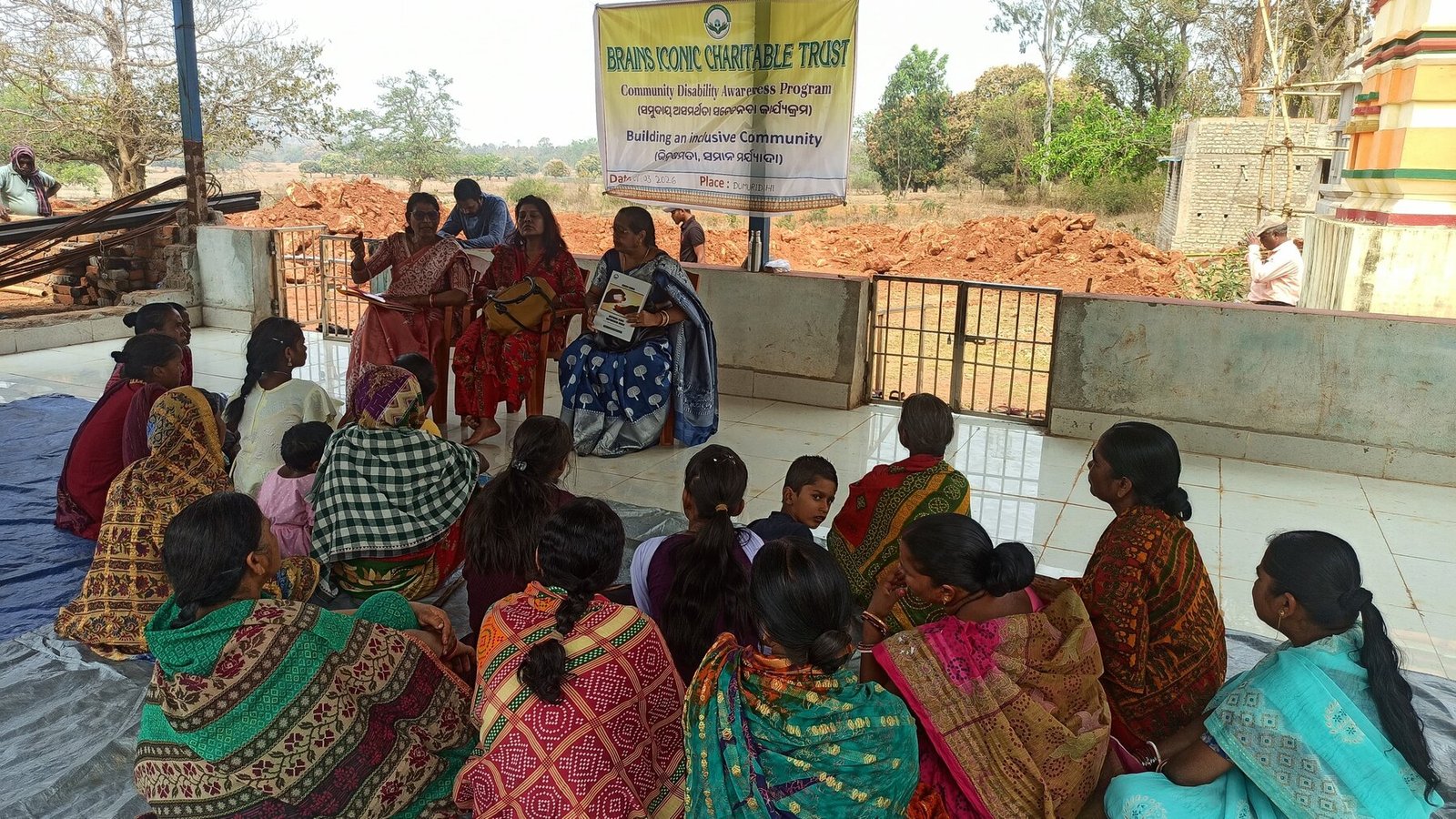 A Brains Iconic Charitable Trust volunteer leads a Community Disability Awareness Program in Dumuridihi, Keonjhar district, seated with women and children under a Building an Inclusive Community banner.
