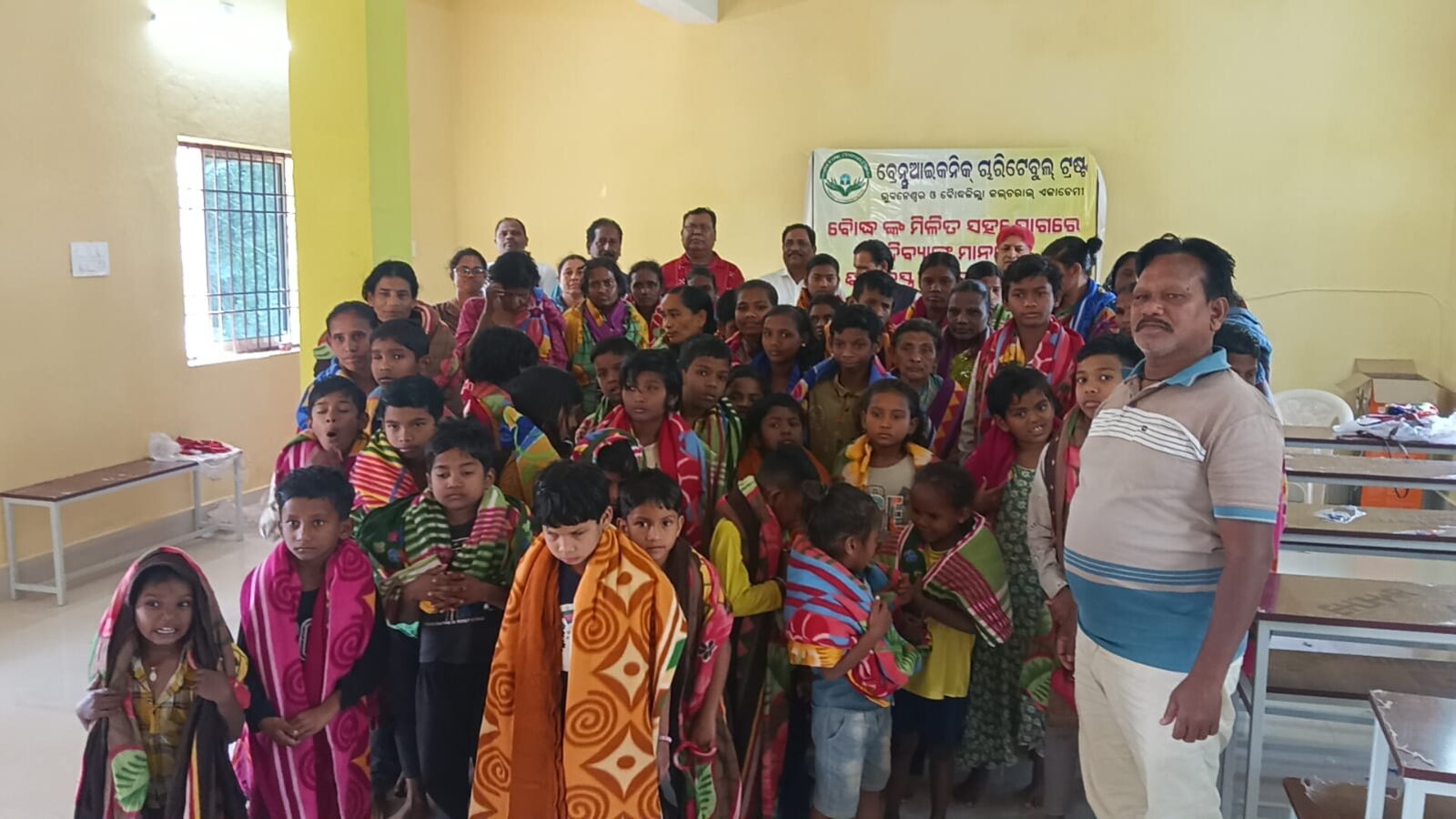 Children from a tribal community wrapped in warm blankets after a Brains Iconic Charitable Trust distribution event, gathered together indoors.